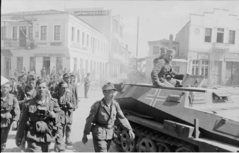 Soldater marscherar på stadsgata. Bredvid militärfordon med två sittande soldater ovanpå.