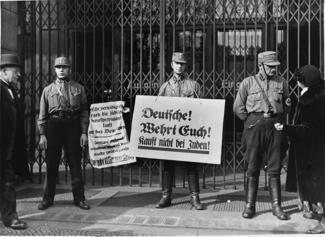 Tre soldater står vakt med plakat med budskapet köp ej av judar.