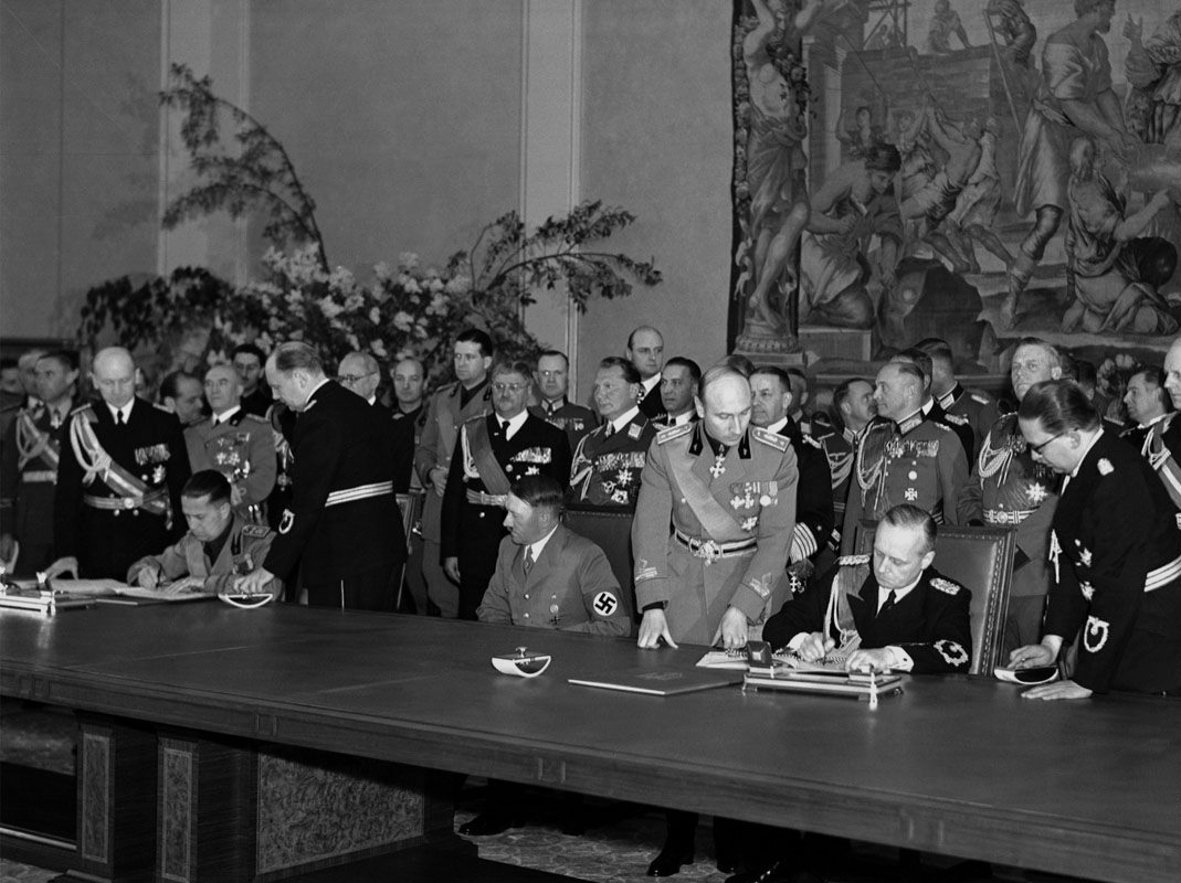 Män signerar dokument vid ett stort bord. Uniformsprydda, dekorerade män i bakgrunden.
