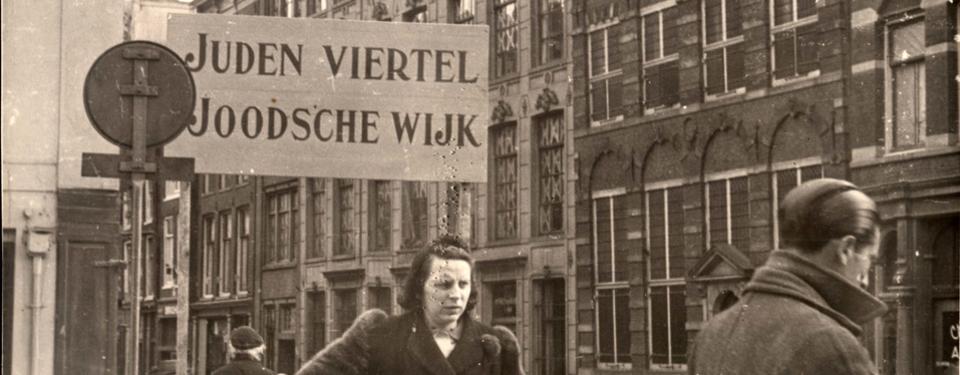 Bilden visar en kvinna och en man på en gata i Amsterdam. På en stor skylt står det: Juden viertel Joodsche wijk.