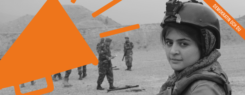 Bilden visar en ung tjej med skyddsutrustning som tittar in i kameran och soldater i bakgrunden.