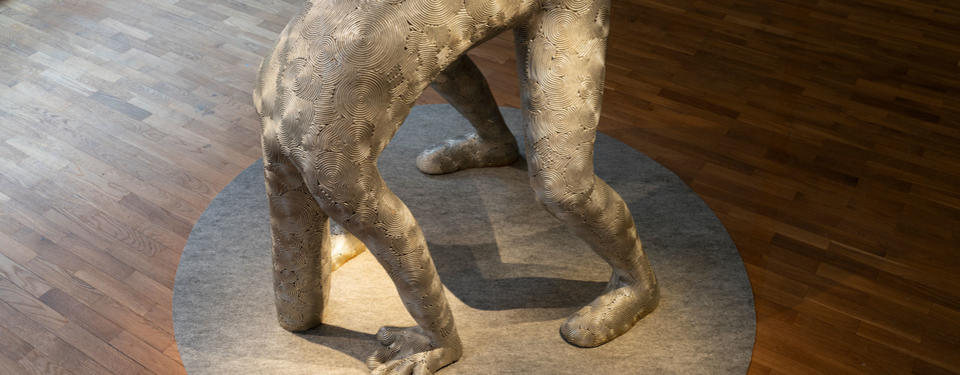Foto på en skulptur som ser ut som en människa. Hen lutar sig framåt och har både fötter och händer i marken. Man ser inte huvudet. Den långa halsen är nerböjd och ser ut att ha stoppat ner huvudet under marken.