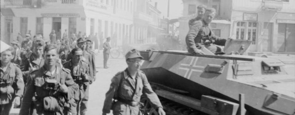 Soldater marscherar på stadsgata. Bredvid militärfordon med två sittande soldater ovanpå.