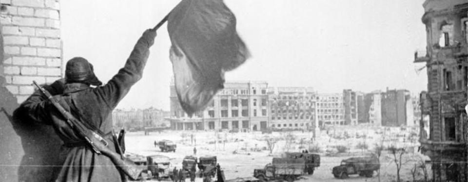 Bilden visar soldat som håller i vit flagg framför stor öppen plats omgärdad av sönderbombade byggnader.
