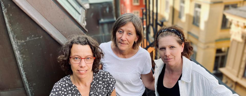 Tre kvinnor står på en takbalkong, i bakgrunden syns takåsarna i Gamla stan.