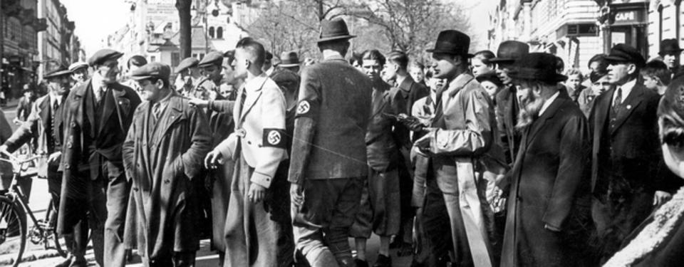 Bilden visar en samling män i kostym på gata, några med armbindel med svastika.