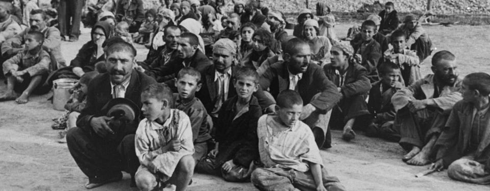Fotografi på en grupp män och pojkar sittandes på marken i ett koncentrationsläger