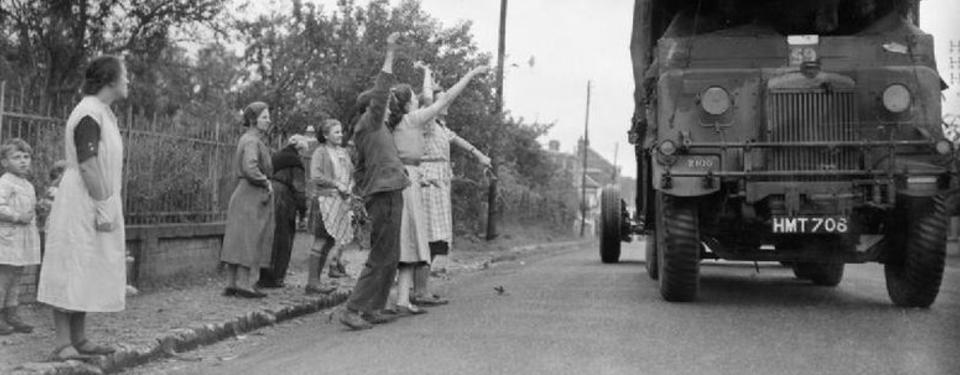 Bilden visar lastbil på gata, sex kvinnor och två barn ståendes vid sidan, varav några vinkar.