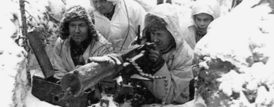 Bilden visar fyra män sittandes i skyttegrav vintertid. En man siktar i vapen.