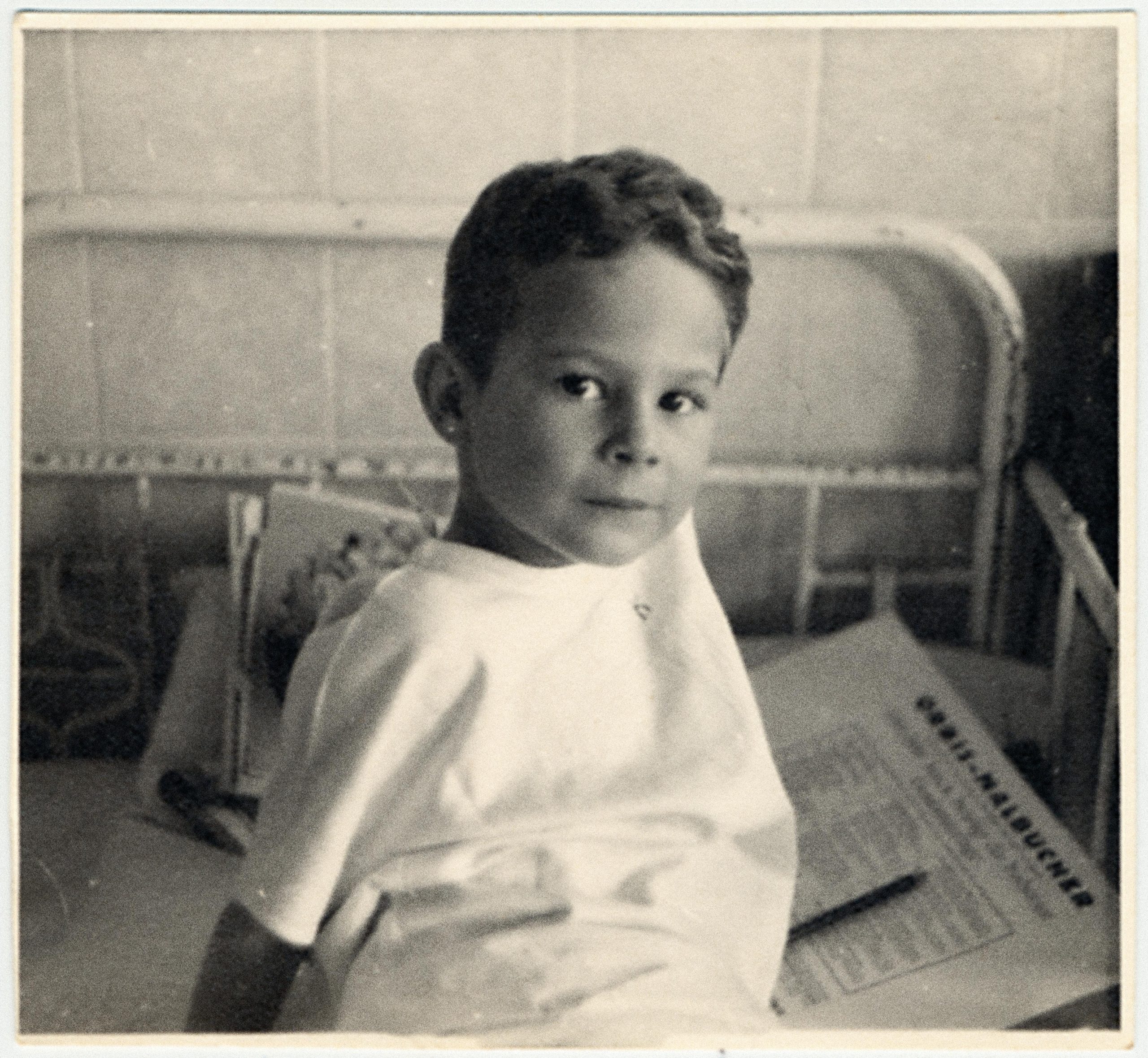 Bilden visar en ung pojke som sitter upp i en sjukhussäng och tittar in i kameran. Det är en äldre säng med stålram och i den finns ett papper och en penna.