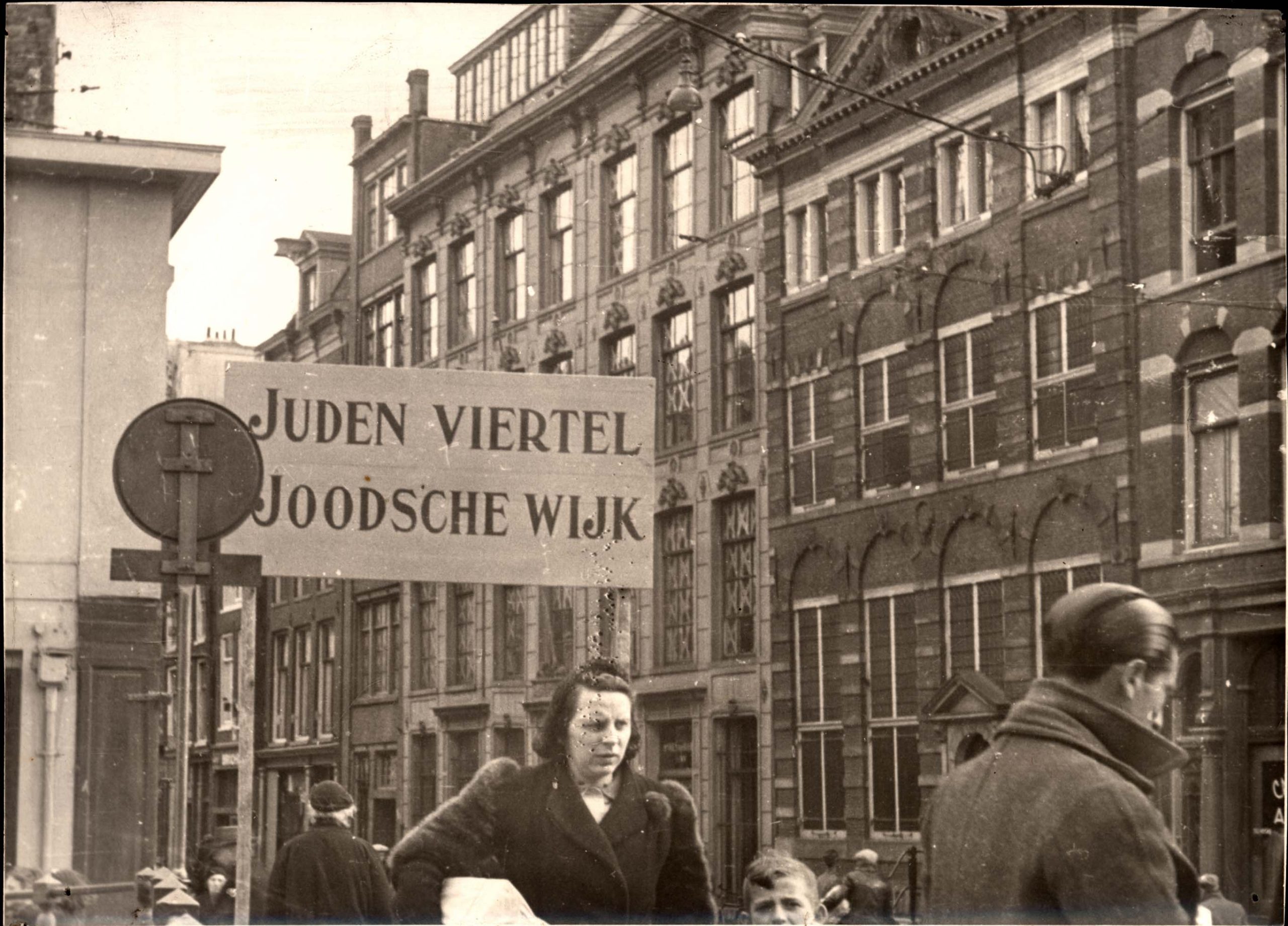 Bilden visar en kvinna och en man på en gata i Amsterdam. På en stor skylt står det: Juden viertel Joodsche wijk.
