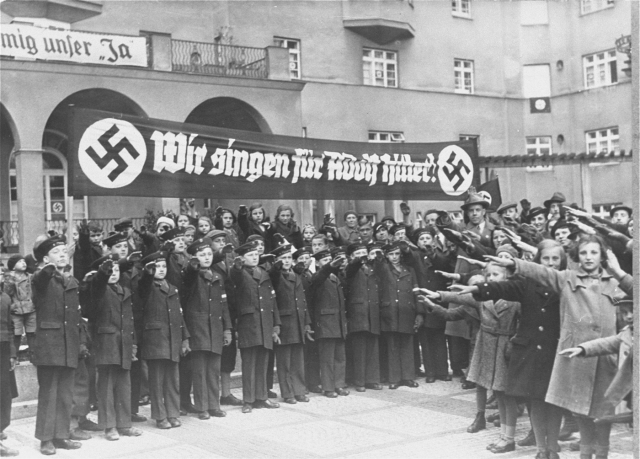 Bilden visar uniformsklädda, hitlerhälsande ungdomar under banderoll med texten wir singen für Adolf Hitler. Vid sidan, civilklädda hitlerhälsande ungdomar.