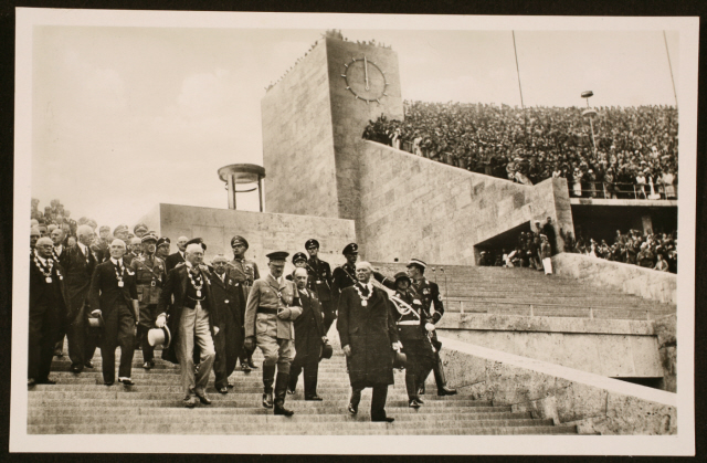 Bilden visar Adolf Hitler omgiven av soldater och välklädda dekorerade civila män gåendes nedför trappa.