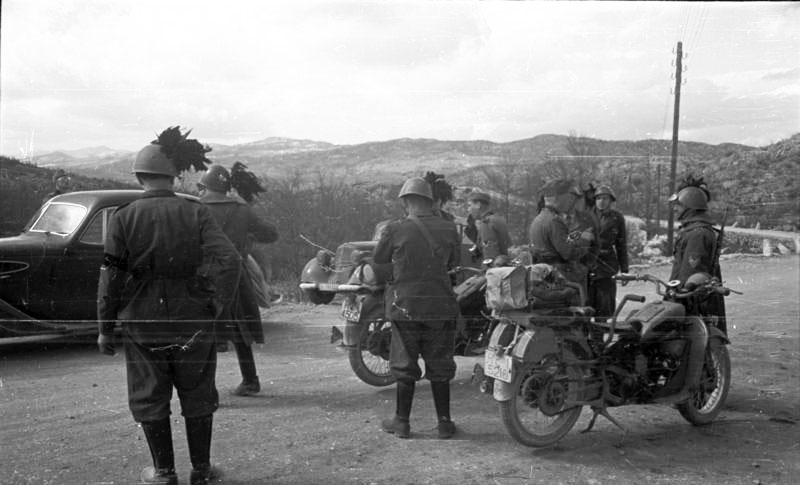 Bilden visar ett tiotal soldater ståendes bland två motorcyklar och två bilar, berg i bakgrunden.