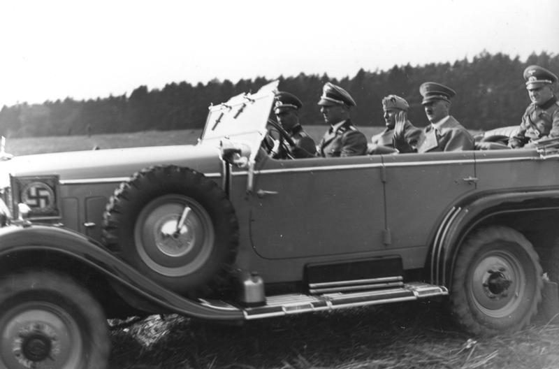 Bilden visar fem män sittandes i öppen bil. Framskärmen dekorerad med en svastika.