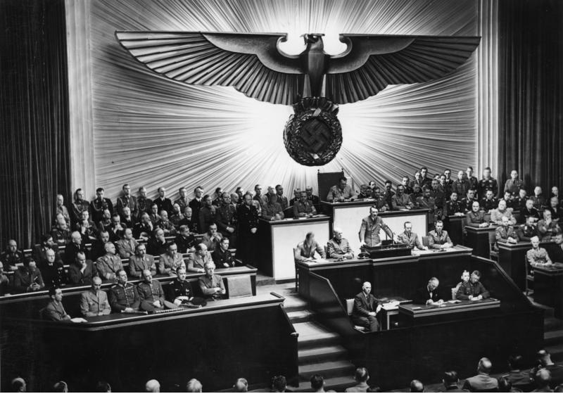 Bilden visar Adolf Hitler ståendes i en talarstol, flera män är bänkade runtomkring honom och i bakgrunden en stor swastika som väggdekoration.