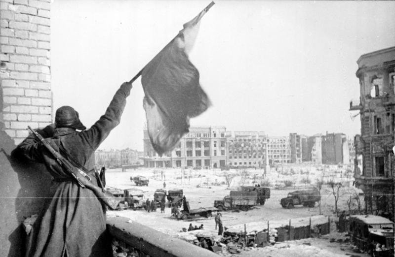 Bilden visar soldat som håller i vit flagg framför stor öppen plats omgärdad av sönderbombade byggnader.