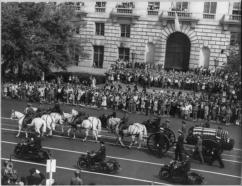 Bilden visar hästdragen vagn med kista täckt av amerikanska flaggan. Flankeras av uniformerade motorcyklister. Folkmassor vid sidan av vägen.