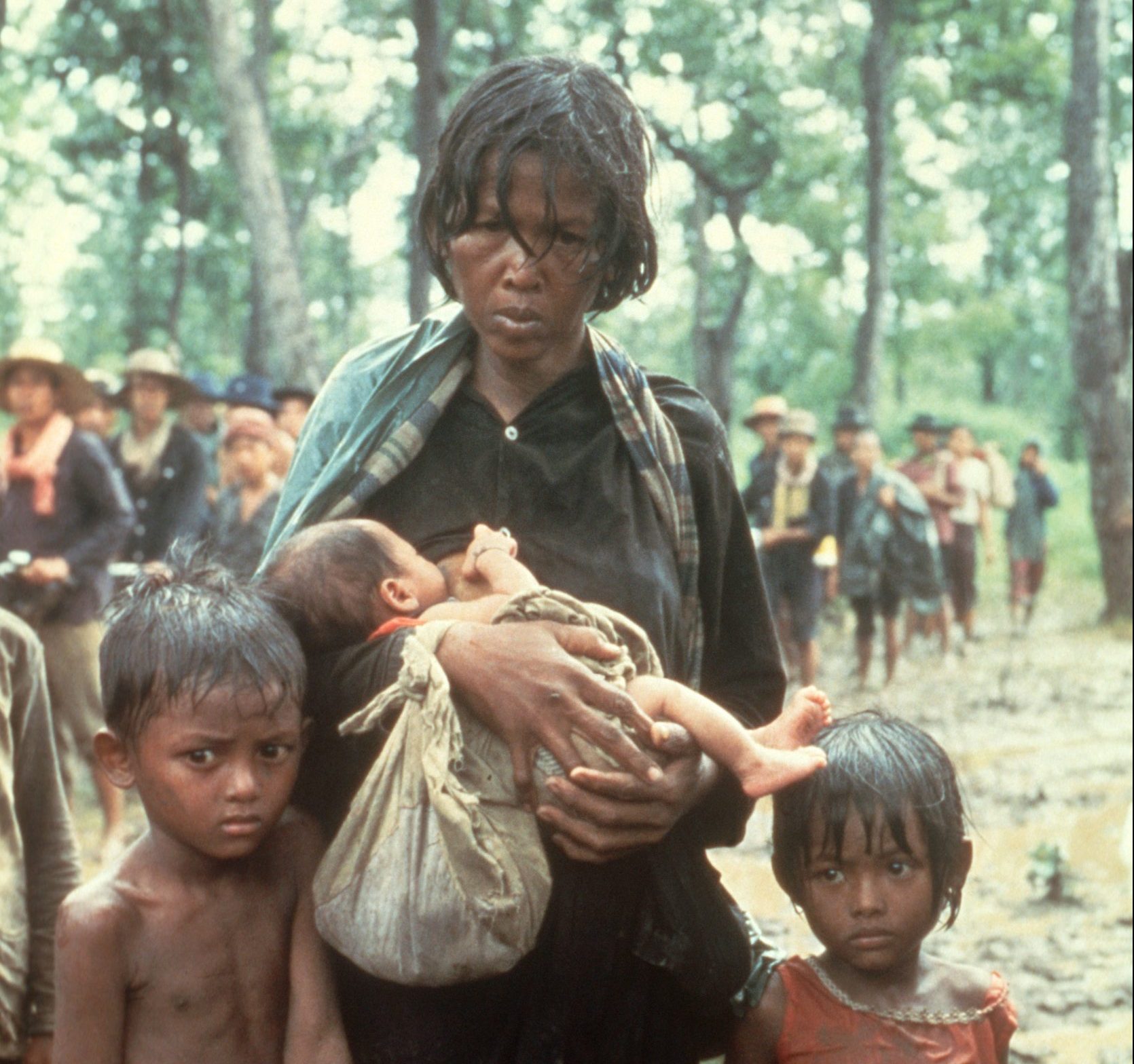 Kvinna i en skog som bär en bebis i sina armar, intill henne står två små barn.