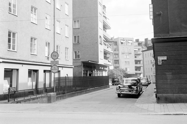 En svartvit bild av en stadsgata . Bilar av 60-talsmodell står parkerade.