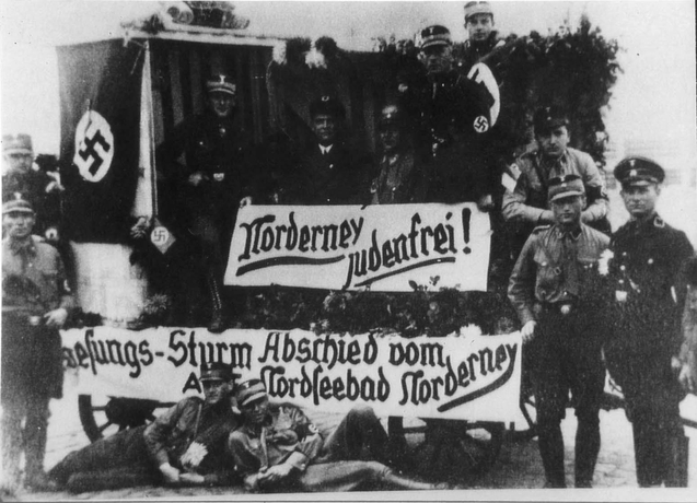 Tre soldater uppställda på sidan av en större vagn med banderollen Norderney judenfrei! Två soldater ligger poserandes framför.