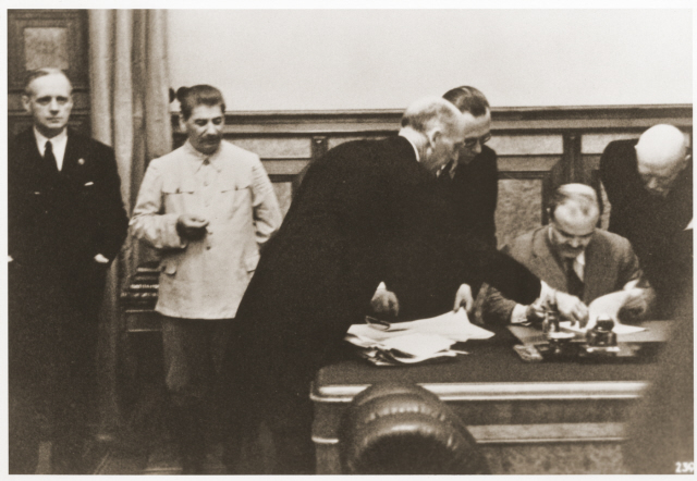 Bilden visar tre män som böjer sig fram mot bord där en man sitter med dokument framför sig.