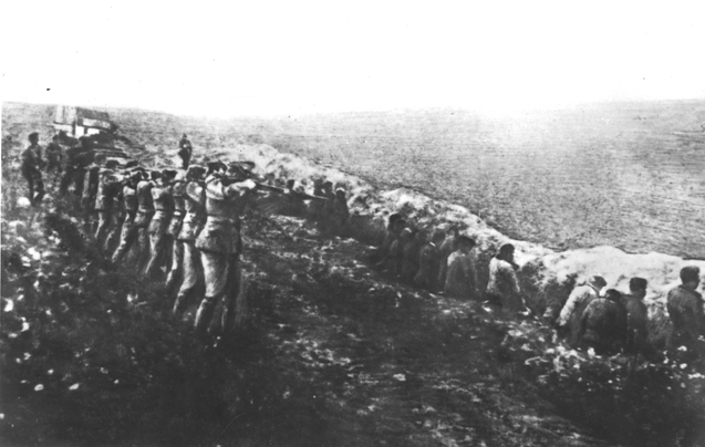Bilden visar ett femtontal soldater som siktar gevären mot uppradade civila med ryggen åt soldaterna, ståendes ett par meter bort.