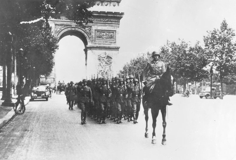 Bilden visar marscherande soldater i tre led med triumfbågen i bakgrunden.