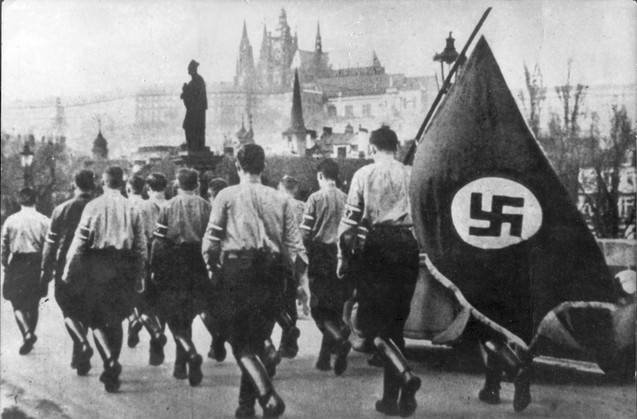 Bilden visar ryggarna på ett tiotal yngre män i nazistuniform, varav en person bär på en fana med en svastika på.