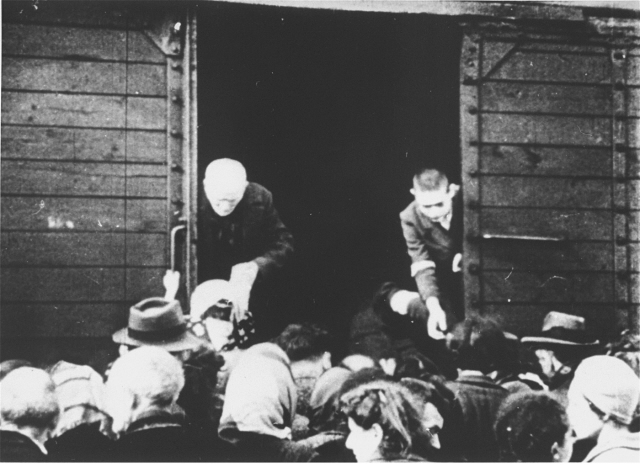 Bilden visar en äldre och en yngre man ståendes i öppningen av tågvagn med många människor utanför.