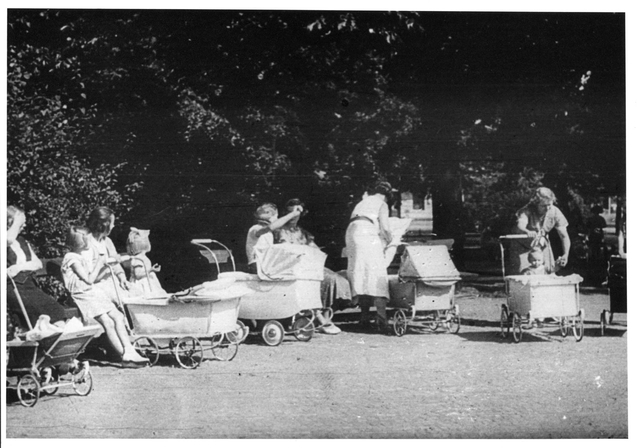 Bilden visar fem barnvagnar i en park med kvinnor och barn i närheten.