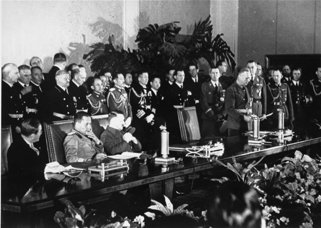Bilden visar tre sittande män vid långt bord, en fjärde man står och läser dokument. Flera kostymklädda, dekorerade män i bakgrunden.