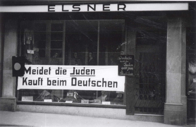 Bilden visar ett butiksfönster till hälften täckt av banderoll med texten: Meidet die Juden Kauft beim Deutschen.
