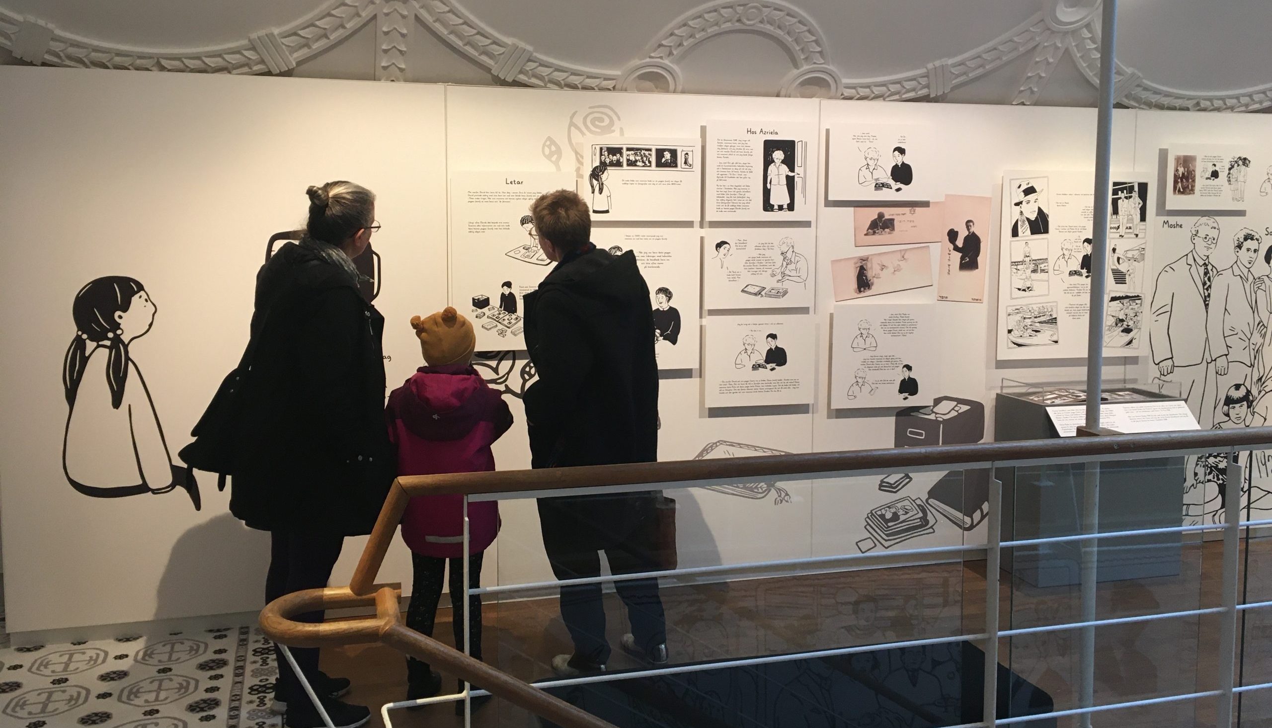 Två vuxna och ett barn tittar på bilder i en utställning.