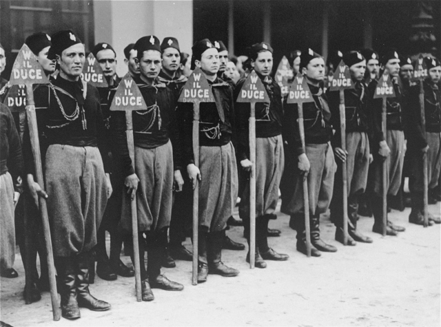 Bilden visar yngre uniformsklädda män på rad i ett par led, hållandes i varsin triangelformad skylt med texten w il duce.