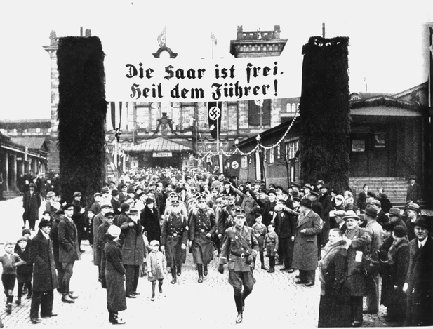 Bilden visar soldater marscherandes i stadsbild omgivna av människor. Banderoll med text Die saar ist frei. Heil dem Führer syns.