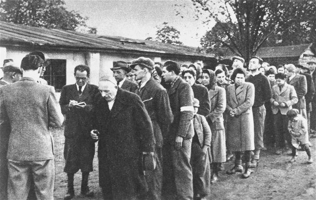 Bilden visar män, kvinnor och enstaka barn ståendes utomhus på led framför man med anteckningsbok.