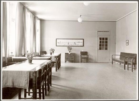 Dagsal på Vipeholms sjukhus. I rummet finns till vänster bredvid fönstren två dukade bord med blommor. Till höger står två bänkar längs väggen. Rakt fram i bilden finns ett bord med blommor och en stol. En tavla hänger ovanför.