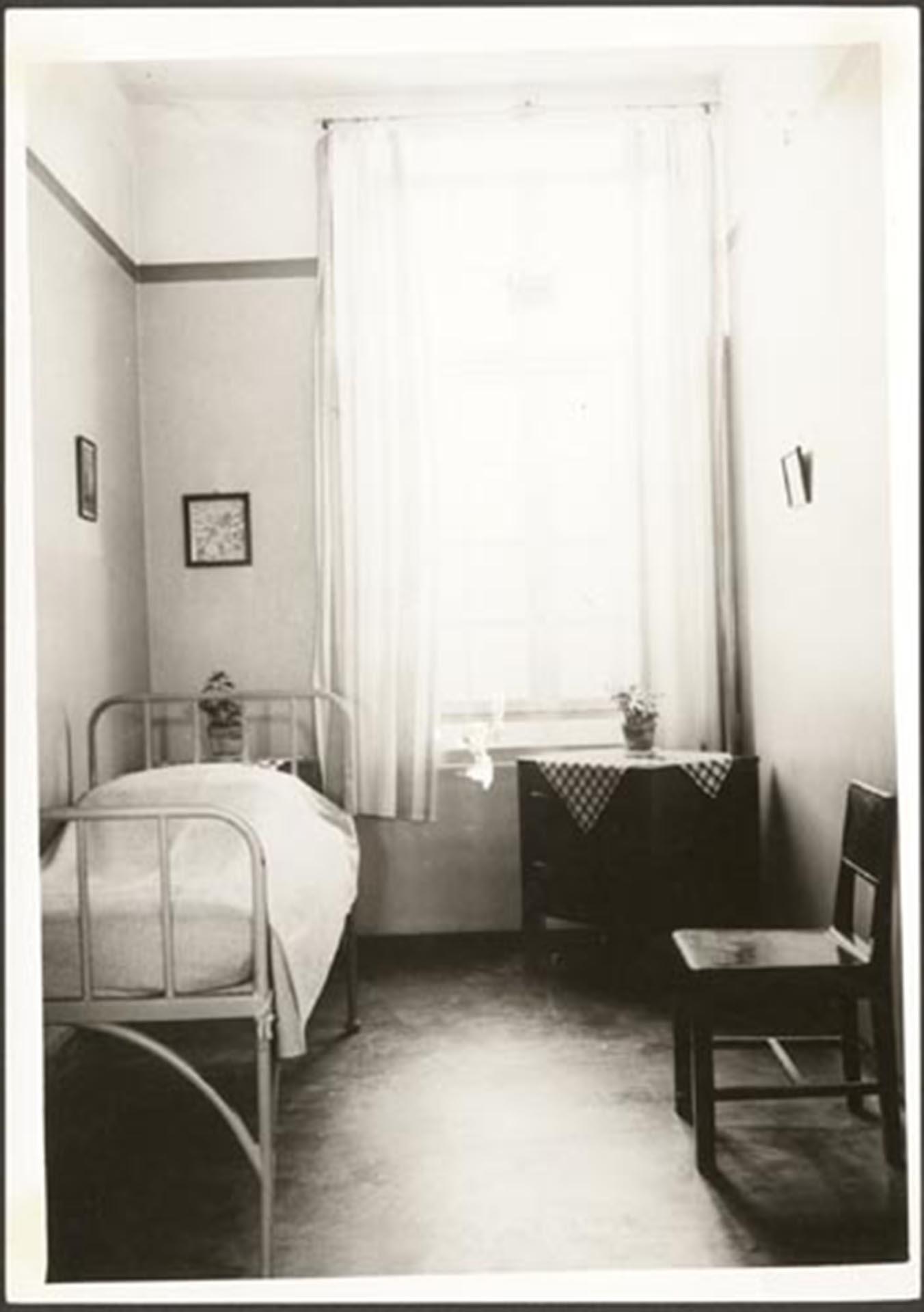 Internrum på Vipeholms sjukhus med en säng, byrå och en stol. Fönster med gardiner och tre mindre tavlor.