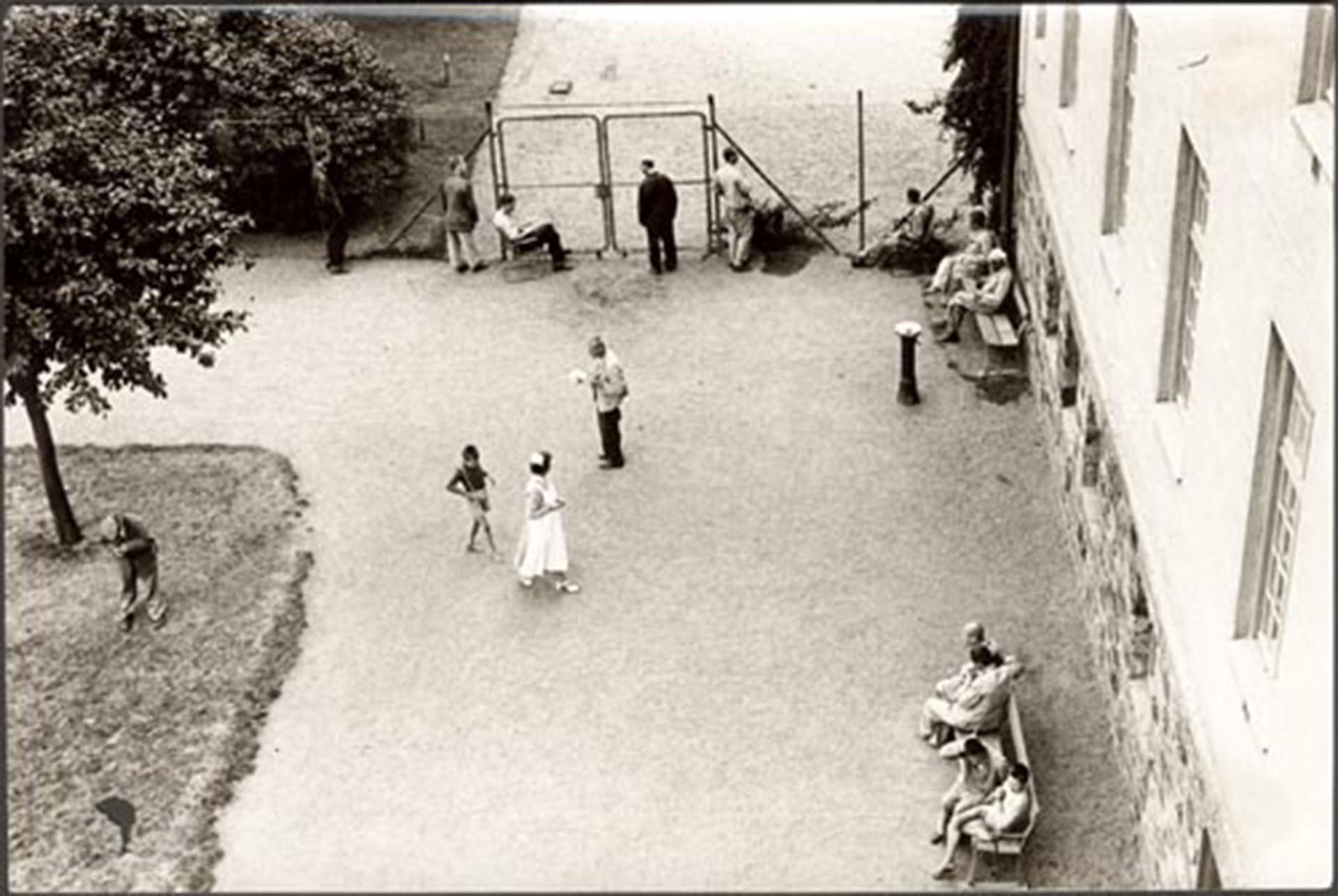 Rastgården på Vipeholms sjukhus snett från ovan, 1953. I mitten av fotot går en patient, troligen ett barn, bakom en anställd i sjuksköterskeuniform. Vid ett staket står några patienter och tittar ut. På två bänkar längs en husvägg sitter några patienter.