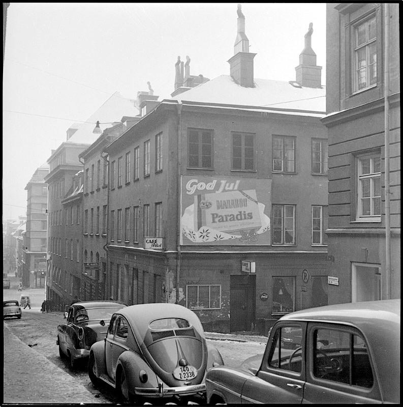 En svartvit bild av en stadsgata på 60-talet. Längst gatan står parkerade tidstypiska bilar.