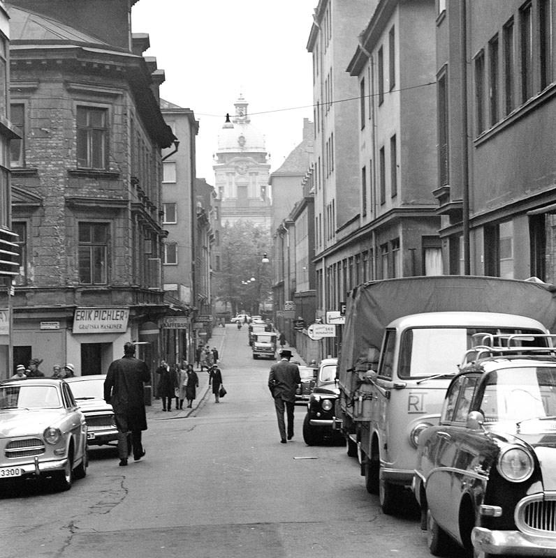 En svartvit bild av en stadsgata på 50-talet. Längst gatan står parkerade tidstypiska bilar. Mitt i gatan promenerar människor.