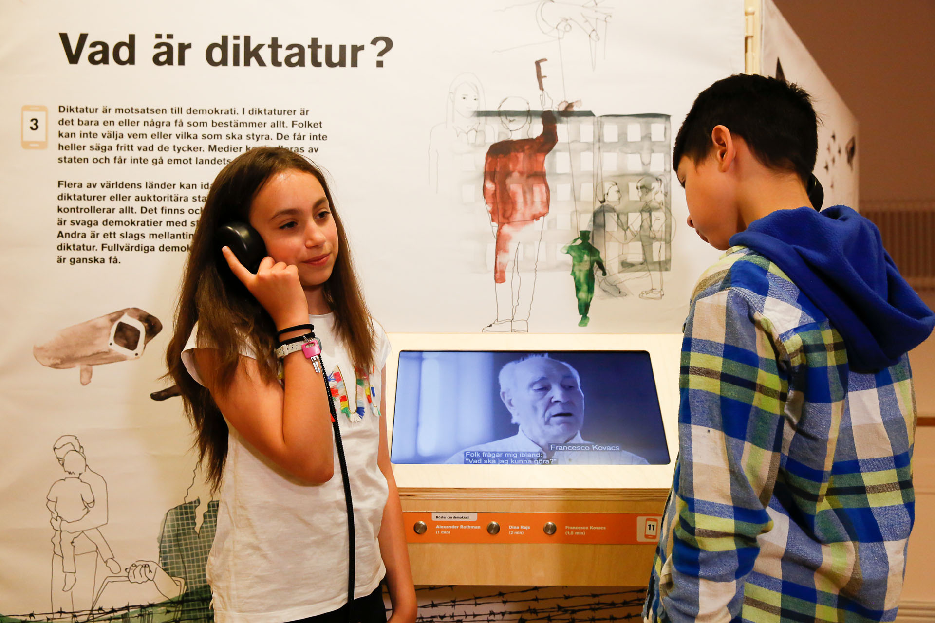 Två barn i tioårsåldern i en utställning. Det ena barnet lyssnar på något i en hörlur och det andra barnet tittar på en digitalskärm som visar en äldre man.