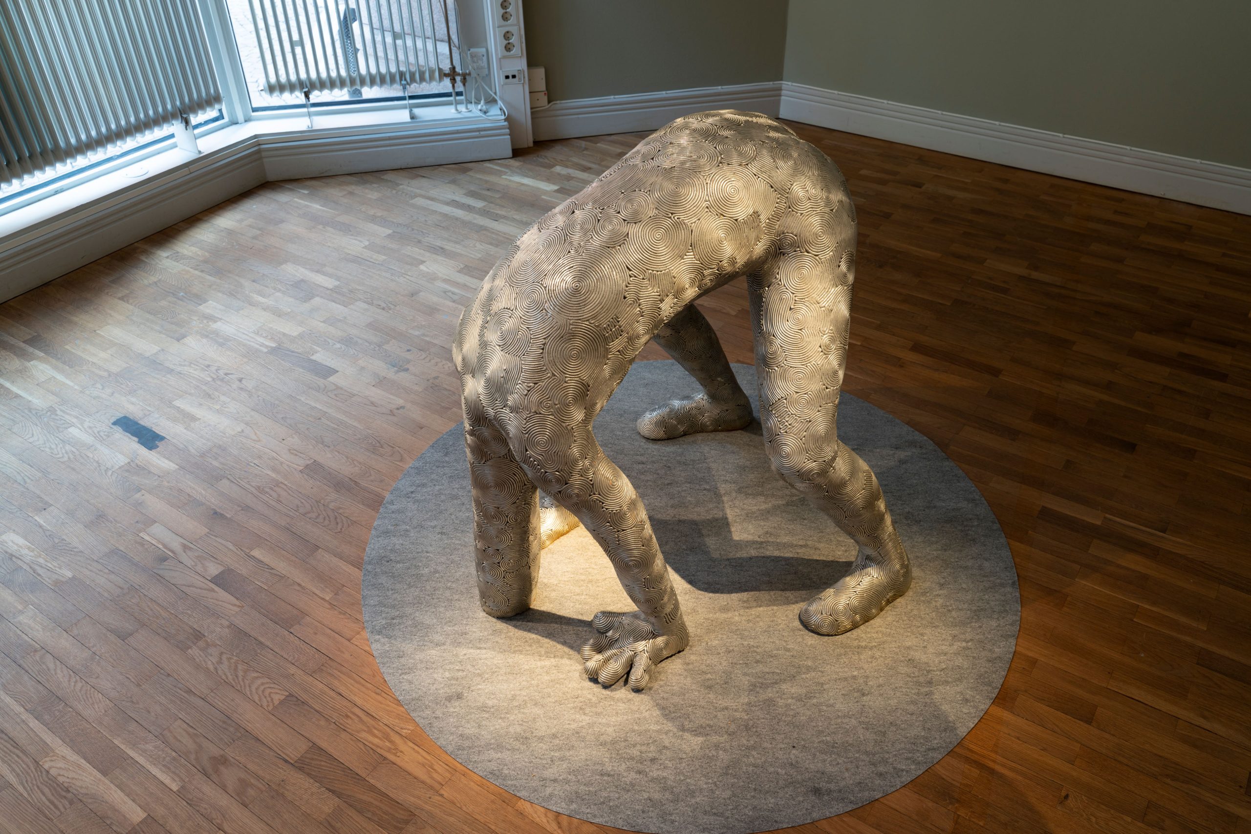 Foto på en skulptur som ser ut som en människa. Hen lutar sig framåt och har både fötter och händer i marken. Man ser inte huvudet. Den långa halsen är nerböjd och ser ut att ha stoppat ner huvudet under marken.