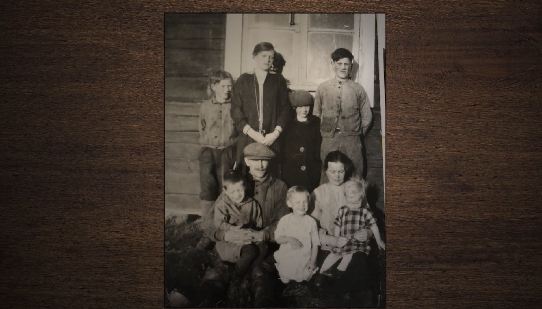Svartvitt äldre foto av en grupp på åtta stycken människor i olika åldrar framför ett hus. Eventuellt är det ett familjefoto.