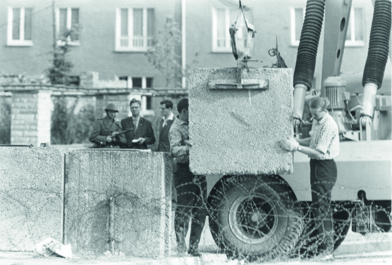 Två personer monterar med hjälp av en kranbil ett betongblock vid byggandet av Berlinmuren.