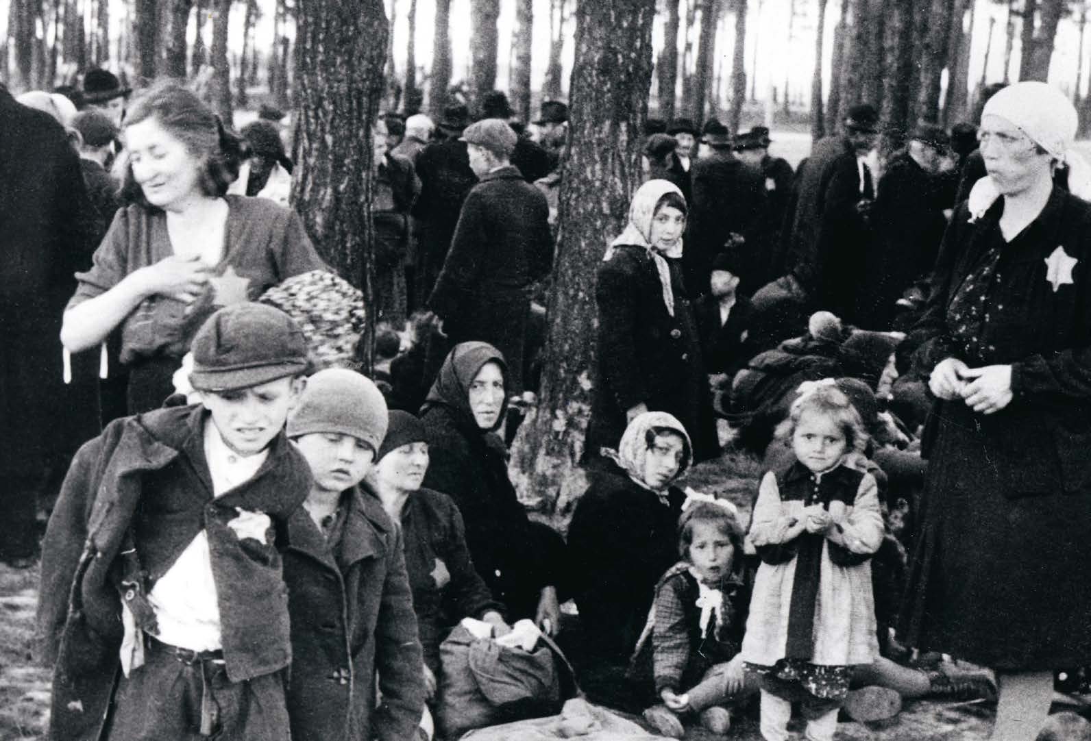 En äldre svartvit bild föreställlande en grupp vuxna och barn som befinner sig i ett skogsområde. Alla ser allvarliga ut.