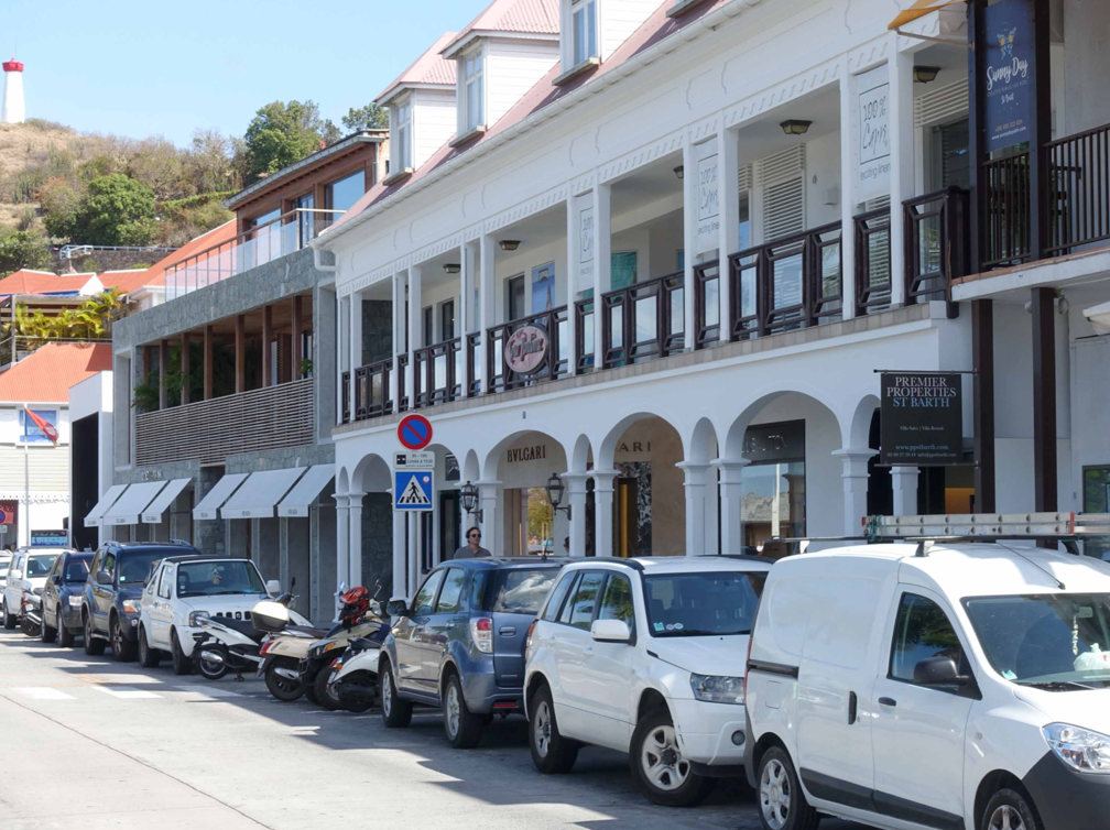 En gata med flera bilar parkerade bilar utanför ett tvåvåningshus. I bildens övre vänstra hörn syns ett vitt fyrtorn.