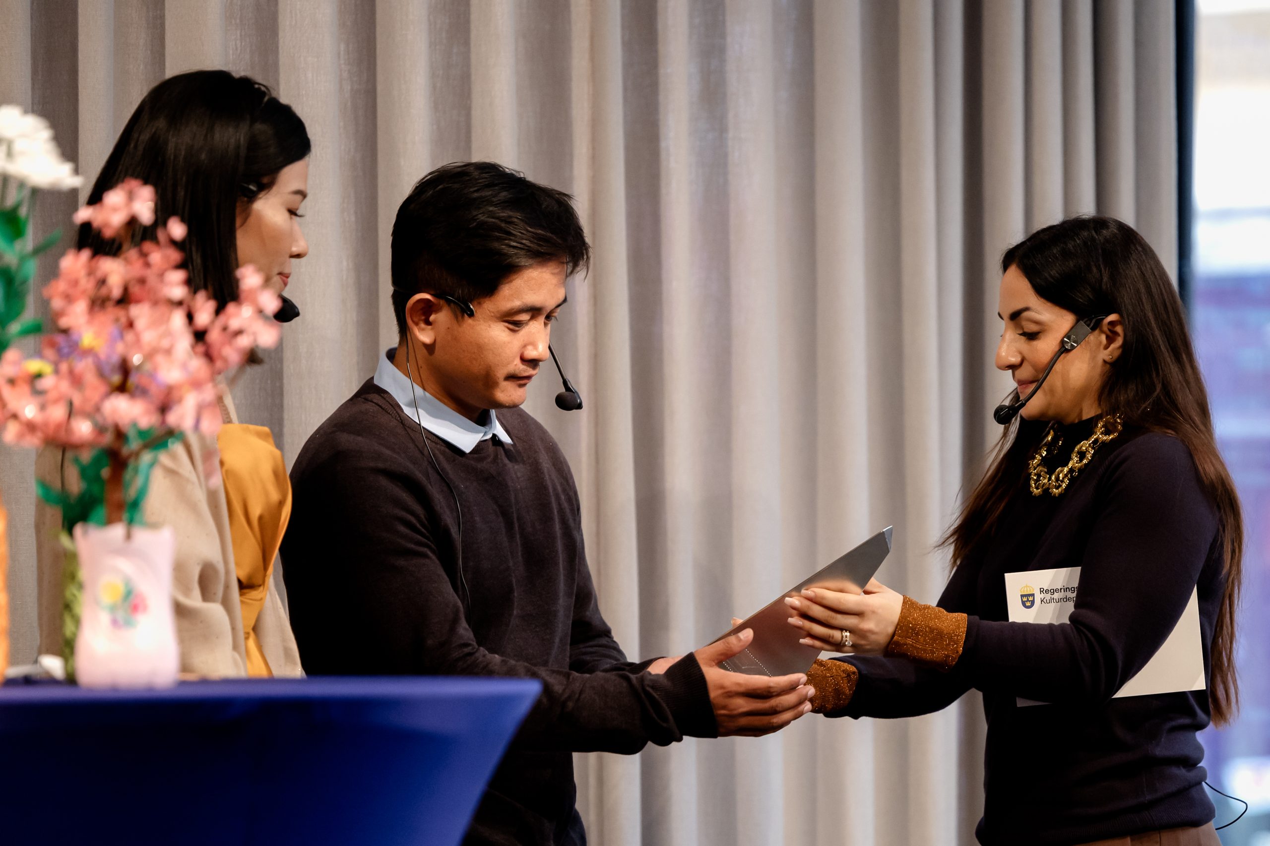 En kvinna räcker över en silvrig prisstatyett till två personer.