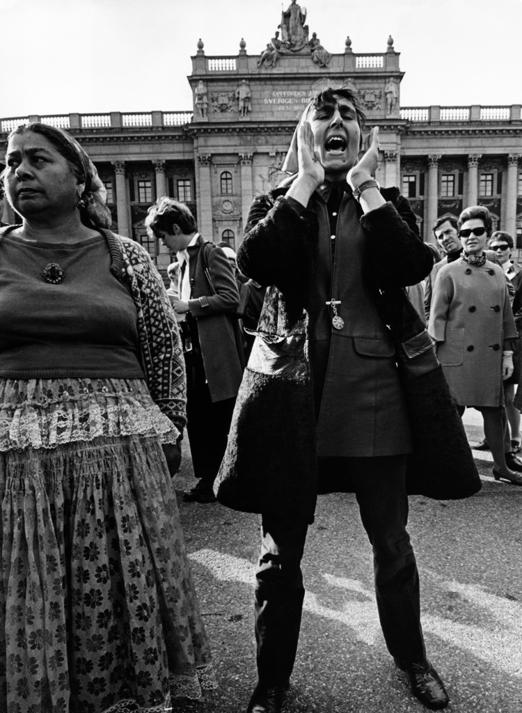Svartvitt foto av en kvinna som står i en folksamling. Kvinnan ropar något och håller händerna som en strut framför munnen för att förstärka hennes röst.